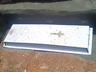 The image shows a white coffin in a grave. The coffin is rectangular with a gold cross affixed to the top. Reddish-brown soil surrounds the coffin on one side, and a darker gray material, likely concrete or similar, lines the other side of the grave. The location, Ninzo South, Nigeria, is based on external knowledge and cannot be confirmed solely from the visual content. There are no people visible in the image.