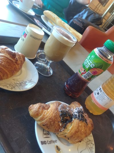 This is a top-down, medium shot photograph taken at a cafe in Livange, Luxembourg, showing a tray with breakfast items. In the foreground, two croissants are presented on white plates with minimalist line drawings of berries and leaves. One croissant, on the right, is topped with white icing and black sprinkles. A bottle of "Fuze Tea" with a green cap is visible to the right of the croissants, with a partially visible bottle of what appears to be juice next to it. A glass mug with a frothy coffee drink, a disposable coffee cup, and a bread roll are also on the tray. In the background, a person's hands are visible, suggesting they are interacting with the items on the tray. The cafe environment is suggested by the brown patterned tray and the presence of other tables and chairs in the blurred background, including a glimpse of orange upholstery. The lighting is bright, possibly from natural daylight, creating reflections on the glass mug and the bottles.