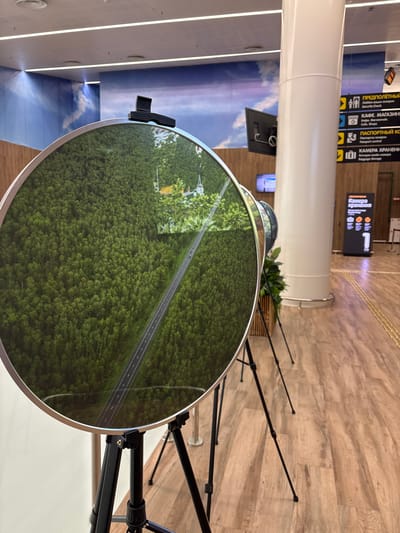 This media file captures an interior scene, likely an airport terminal or a public transit station, given the directional signage. The main subject is a large, circular reflective surface that displays a vibrant aerial view of a dense green forest bisected by a grey asphalt road. The reflection suggests this is a display or a decorative element.

In the background, typical airport elements are visible. To the left, a large white pillar dominates the frame, with a digital display or mural depicting a sky with clouds on the wall behind it. Above and to the left of the circular display, directional signs in Cyrillic script are mounted on a wall, indicating "ПРЕДПОЛЁТНЫЙ" (Pre-flight), "КАФЕ. МАГАЗИНЕ" (Cafe. Shops), "Пастерная аскарен" (Passport control), and "КАМЕРА ХРАНЕНИ" (Baggage Storage). These signs, along with the modern lighting and polished floor, confirm the setting is a transportation hub.

The circular display is supported by a black tripod structure, implying it might be a temporary or portable exhibit. A small black clip is attached to the top edge of the circular display. A potted green plant is visible behind the display, adding a touch of nature to the otherwise man-made environment. The lighting appears to be artificial, suggesting an indoor environment, likely during the day. There are no people visible in the frame, emphasizing the objects and the setting. The overall impression is one of modern public space with a visually interesting display.