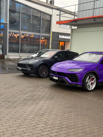 The image shows two high-end sports cars parked on a wet, paved lot in front of a building with a sign that reads "VISAGE." The car in the foreground is a bright purple Lamborghini Urus, and behind it is a dark blue Porsche Macan. The building appears to be a commercial establishment, possibly a club or restaurant, with large glass windows and interior lighting visible. The sky is overcast, and the ground is wet, suggesting it has recently rained or is raining. The license plate on the Lamborghini is visible and reads "ZH 31194U." The image captures the cars and the building facade, with the overall impression being one of modern urban luxury. Dübendorf, Switzerland is known for its mix of residential and commercial areas, and this scene is consistent with a business district or entertainment venue within the city.