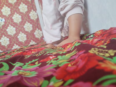 In a dimly lit room, a child's legs and feet are visible, clad in a light-colored pajama set with faint red stains. The child is standing on a bed covered with a burgundy duvet featuring a vibrant floral pattern of red, pink, green, and yellow flowers and leaves. To the left, a portion of a wall with red wallpaper adorned with an intricate damask pattern of cream and gold motifs is visible. The overall scene appears to be indoors, likely a bedroom, with no clear indication of the specific location within Социализм, Kyrgyzstan.