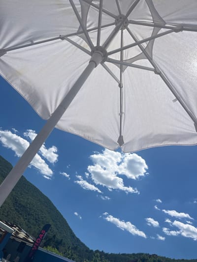 The image was taken at the "VIDAS" beach bar in Varvara, Bulgaria. It is a low-angle shot looking up at a white beach umbrella with a clear blue sky and scattered white clouds in the background. The lush green slopes of a mountain can be seen on the left side of the frame, with trees and part of a building structure visible at the bottom. The umbrella is open and appears to be made of fabric stretched over a metal frame. The lighting suggests it is a sunny day.