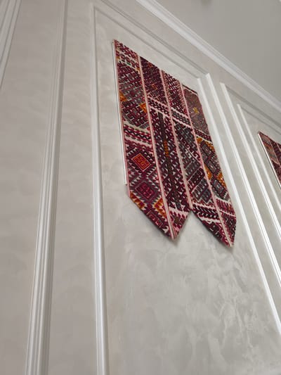 A low-angle, indoor shot shows a white, textured wall with raised decorative paneling. Two pieces of intricately patterned fabric, primarily red, white, and gold, are hung diagonally on the wall. The fabric pieces are triangular in shape with decorative geometric patterns. To the right, a portion of another similar fabric piece is visible. The scene appears to be a residential or commercial interior space. The location is identified as Zhanaozen, Kazakhstan.
