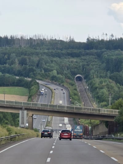 The image shows an aerial view of a highway and a train tunnel through a forested hillside, likely in Deesen, Germany. The highway is paved with asphalt and marked with white lane lines. Several vehicles are on the highway, including cars, trucks, and vans. One truck is blue and has the text "AE EBNER" printed on its side, along with a logo. Another truck is silver, and a red car is visible. The train tunnel is curved and appears to be made of concrete. Train tracks lead into and out of the tunnel. The surrounding area is covered in dense green trees, with some bare trees visible on the upper part of the hill. The sky is overcast with some clouds. The overall scene depicts a transportation infrastructure integrated into a natural landscape. There are no people clearly visible. The time of day appears to be daytime, and the weather seems to be cloudy.