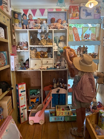 The image depicts the interior of a toy store located in Kfar Vradim, Israel. The store is filled with a wide assortment of toys, primarily dolls and stuffed animals, displayed on wooden shelves. In the foreground, a young girl wearing a straw hat and a pink backpack is holding a large stuffed monkey. She is facing away from the camera, interacting with the toy.
The shelves are organized with various types of toys. On the upper shelves, there are several dolls, including baby dolls and fashion dolls, arranged in groups. Below them, a collection of stuffed animals, such as bears, lions, and elephants, are prominently displayed. Some of the shelves also contain boxes of toys and other smaller decorative items.
In the lower part of the image, there are wooden toy pushcarts, building blocks with numbers, and toy trucks. The store's walls and shelves are made of wood, contributing to a rustic and charming atmosphere. Natural light appears to be entering the store from an unseen source, illuminating the display. The overall scene suggests a well-stocked and inviting toy shop.