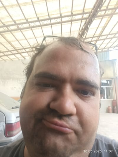 The image is a close-up, selfie-style photograph of a man's face, taken outdoors under a corrugated metal roof structure. The man is sweating, with visible moisture on his forehead and hairline. His mouth is pursed, and he appears to be looking directly at the camera. In the background, parts of a silver car and a building with windows are visible. The overall lighting is bright, suggesting daylight. The date stamp "30.06.2026 14:07" is overlaid on the bottom right of the image. There is no information in the image to suggest the location is the city of Petrovka, Kyrgyzstan.