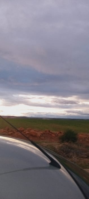 A tilted, slightly blurry view of a landscape from inside a vehicle. The sky fills the left, with pale blue merging into bands of white clouds illuminated from behind. A strip of green vegetation borders a reddish-brown terrain to the right. Part of a white vehicle interior, possibly the roof or hood, fills the lower right corner of the frame.