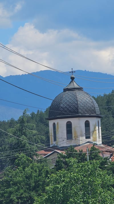 This is a vertical, medium shot of a church dome and part of its structure, set against a backdrop of trees and mountains. The dome is dark gray, with a diamond-patterned surface, and topped with a small spire and cross. The church building is light-colored with arched windows. Several electrical wires crisscross the foreground, obscuring parts of the scene. Lush green trees are visible on the right and behind the church. In the background, layered mountains fade into a blue sky with scattered white clouds. The lighting suggests it is daytime, likely a sunny day. The scene appears to be in a rural or semi-rural mountainous area, consistent with the provided location context of Slatina, Bulgaria. There is no discernible activity or event taking place, and no text is visible.