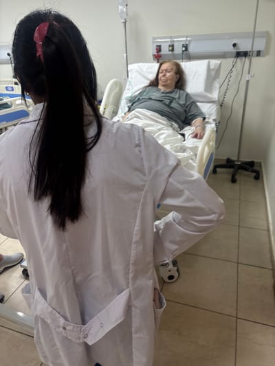 The image depicts an interior scene within a medical facility in Adrogué, Argentina. In the foreground, a female medical professional is viewed from behind, standing and facing an elderly female patient. The professional has long, dark hair secured with a pink claw clip and wears a white lab coat over light-colored attire, with a light-colored sneaker visible on the left.

The patient, appearing to be between 60-80 years old with reddish-brown hair, lies supine in a hospital bed with white bedding. She wears a grey short-sleeved top, and her eyes are closed. An intravenous (IV) line is inserted into her left forearm (viewer's right), and a nasal cannula is present, delivering oxygen. An IV bag hangs from a chrome pole positioned to the right of the patient's head. A medical headwall unit with electrical outlets and service ports is mounted on the light-colored wall above the patient's bed. The room features light-tiled flooring and is illuminated by bright overhead lighting. The medical professional is positioned facing the patient.