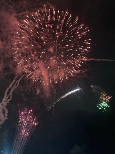 Fireworks explode in a dark night sky. A large burst of red and white fireworks dominates the left side. Above it, smaller bursts of green and yellow are visible. To the right, a silver streak descends, followed by a cluster of red sparks trailing downwards. Smoke from the explosions lingers in the air, creating a hazy atmosphere.