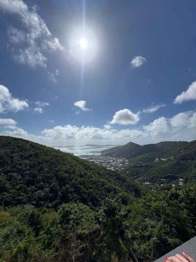 A scenic view of a lush, green mountainside, speckled with buildings, meeting a shimmering body of water. A bright sun shines in a partly cloudy blue sky, with its light reflecting on the water. The trees form a dense, tropical forest covering the hillsides. The image is slightly tilted, giving a dynamic perspective.