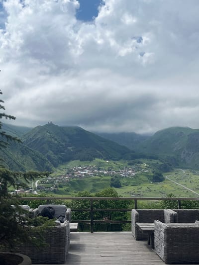 A medium shot captures a scenic view from an outdoor seating area in Stepantsminda, Georgia, looking out over the valley. In the foreground, a wooden deck is furnished with grey wicker seating and a wooden table, with a large pine tree branch partially obscuring the left side. In the mid-ground, a village of houses with red roofs is nestled in a lush green valley, with a winding road visible. In the background, rolling green mountains rise under a cloudy sky, with a prominent church structure perched atop one of the peaks. The scene is peaceful and serene, with a sense of vastness and natural beauty.