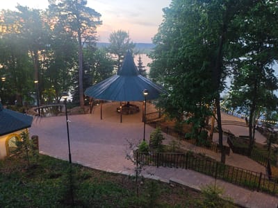 The image depicts a public park in Krasny Klyuch, Russia, taken at dusk. A large, gazebo-like structure with a conical, dark-green roof dominates the center of the scene. The structure sits on a brick-paved area. Inside the gazebo, benches and tables are visible, illuminated by warm lighting. Streetlights also surround the gazebo, adding to the ambient light. To the right of the gazebo, a series of brick staircases with metal railings descend towards what appears to be a body of water, likely a river or lake, which is partially visible in the background. The water's edge is adorned with string lights. Trees frame the gazebo and the staircases, creating a natural border. To the left of the gazebo, a small, illuminated building with an arched window and green-tiled roof is visible. The foreground is a slightly elevated, grassy area with small trees and shrubs. The overall atmosphere is tranquil and inviting. The setting sun casts a soft, warm glow on the scene. There are no people visible in the park from this aerial perspective, likely captured by a drone.