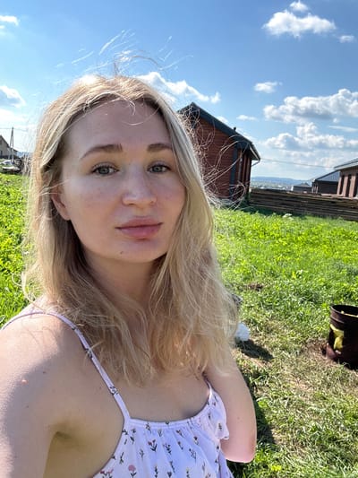 A young Caucasian woman with blonde hair and fair skin is taking a selfie outdoors on a sunny day. She is wearing a light-colored dress with a floral pattern. Her eyes are green, and she has a mole on her cheek. The background shows a grassy hill with several wooden structures, likely houses or sheds, and a stack of lumber. The sky is blue with scattered white clouds. The location is identified as Mokino, Russia, implying a rural or semi-rural setting.
