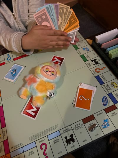 The image depicts an indoor scene in Gerakas Municipal Unit, Greece, featuring a Monopoly board game in progress. A pair of hands, belonging to an individual wearing a light-colored zippered hoodie, occupies the upper left portion of the frame. These hands are fanned out, holding various denominations of Monopoly currency, including light blue, pink, purple, green, beige, orange, and gray notes, with "MONOPOLY" visible on some. Centrally positioned on the board is a multi-colored plush toy, predominantly yellow, orange, light blue, and white, featuring a distinct face with large eyes exhibiting rainbow irises and a wide smile. A small white die, showing faces 2 and 3, rests near the plush toy's right paw.
The game board, with text primarily in Greek, is laid out on a flat surface. To the left of the plush toy, a stack of blue game cards is visible. To its right, an adjacent stack of orange cards with a white question mark is present. A silver-colored car-shaped game token is positioned on a brown property square labeled "ΟΔΟΣ ΕΠΤΑΝΗΣΟΥ" (Eptanisou Street). Portions of a black plastic game organizer tray, containing stacks of cards and money, are visible in the upper right corner. The board displays copyright information including "1935, 2021" and the "HASBRO Gaming" logo. The scene suggests a recreational activity involving the board game.