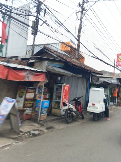 A street scene shows an outdoor shop with goods displayed. Two motorcycles are parked nearby, one with a white delivery box. A person is riding one of the motorcycles. Above the shop, a network of power lines obscures the sky. Additional buildings and signage are visible in the background.