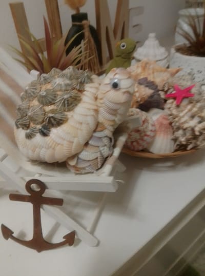 The image depicts a close-up, slightly tilted view of a decorative seashell turtle, which appears to be a craft item, positioned on a miniature white deck chair. The turtle is adorned with various seashells, some of which are arranged to resemble scales, and two googly eyes. Behind the turtle, there's a collection of seashells in a shallow dish, a small green toy lizard with a googly eye, and a pink plastic starfish. In the background, wooden letters spelling "HOME" are visible, along with a dark bottle with twine. To the left of the turtle, a decorative brown anchor is placed on the surface. The scene is set against a white background, suggesting an indoor display or shelf. There is no discernible indication of the city of Wedel, Germany.