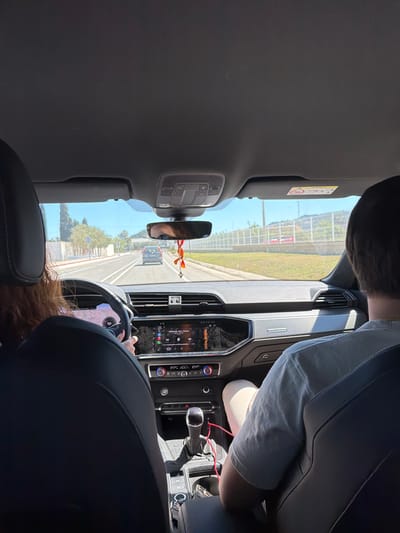 The image shows the interior of a car traveling on a road. The car's dashboard, rearview mirror, and driver are visible. A person is in the passenger seat. Outside, the view includes a road with trees and greenery. The location is likely Beniflà, Spain.