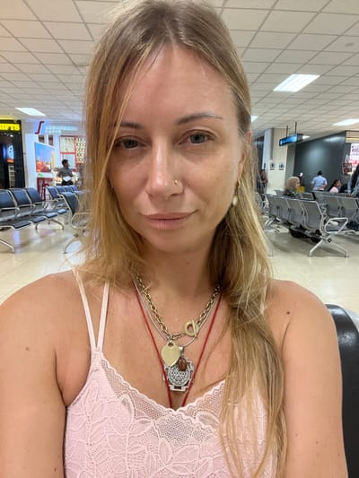 A woman with blonde hair and a light pink lace top is seated in what appears to be an airport waiting area. She is looking at the camera and wearing several necklaces. The background shows rows of empty seats, a cafe, and other people. The environment suggests a public transportation hub, consistent with being in Katunayake, Sri Lanka, home to Bandaranaike International Airport.