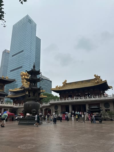 The image depicts the Jing'an Temple in Jing'an District, Shanghai, China, on a cloudy day. The temple's traditional architecture, characterized by golden roofs and ornate details, stands in contrast to the modern glass skyscrapers that surround it. In the foreground, a large, weathered bronze incense burner with intricate carvings and two lion sculptures at its base is centrally positioned. Several individuals are present in the courtyard, some holding umbrellas, indicating potential rain. A group of people are gathered on a raised walkway in front of one of the temple buildings, which features a golden, multi-tiered roof adorned with dragon motifs. Behind the temple, two tall, modern buildings with reflective glass facades dominate the skyline. The ground in the courtyard is paved and appears wet, with circular decorative patterns visible. In the distance, more trees and buildings can be observed. The overall atmosphere is one of juxtaposition between historical and contemporary urban elements.