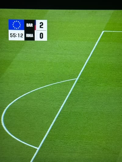 The image shows a green soccer field with white lines marking the penalty area. The scoreboard indicates a match between BAR (Barcelona) and RMA (Real Madrid) with a score of 2-0 at 55:12. The location in Babynino, Russia, is not evident from the image.