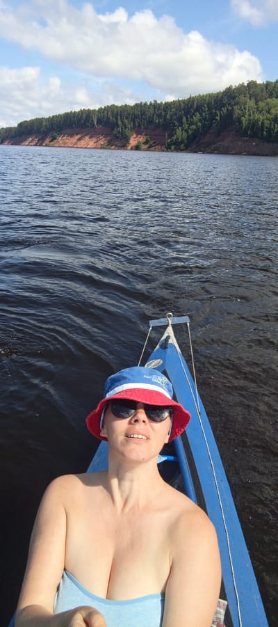 In the city of Kostovaty, Russia, a woman wearing a blue and red bucket hat and sunglasses is pictured from the chest up, seated in a blue boat on a dark, rippling body of water. She is looking towards the viewer with her mouth slightly open. Her top is light blue and strapless, revealing her shoulders and cleavage. The boat's blue hull is visible to her right, and a silver hook is attached to the front of the boat. In the background, a forested cliff face with red earth is visible along the horizon, under a partly cloudy sky. The water is choppy, with some small waves.