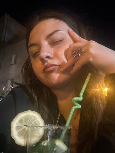 A selfie taken at night shows a young woman with her eyes closed, resting her forehead with her fingers. She is wearing a ring on her right hand. In the foreground, a cocktail glass with a lemon slice and a green straw is visible. The background appears to be an outdoor urban setting with a building and lights. The image seems to have been taken in Noyon, France.