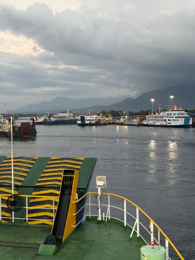 The image depicts a ferry terminal in Melaya, Indonesia, at dusk or dawn. In the foreground, the deck of a ferry is visible, featuring a ramp with yellow and green chevron markings, a black winch, and a green cylindrical object with a red wheel. White railings with a yellow handrail line the edge of the deck. In the middle ground, several ferries are docked at a pier. One prominent white ferry, branded "RLU," is brightly lit. Other ferries, some appearing older and industrial, are also present. The background shows a shoreline with distant mountains under a cloudy, overcast sky. The water is calm, reflecting the lights from the terminal and ferries. The scene suggests a busy port environment, likely a transportation hub for vehicles and passengers.