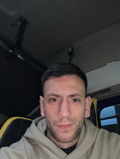 A male individual is depicted in a head-and-shoulders close-up, appearing to be a self-portrait. He has short, dark hair, light stubble on his face, and brown eyes, maintaining a neutral expression directed at the camera. A distinct reddish mark is visible on his forehead, centrally located. He is wearing a light-colored (possibly beige or light grey) hooded sweatshirt over a dark undergarment. The background indicates an interior environment, consistent with a vehicle cabin. To the left, a portion of a black seat with yellow trim is visible. The dark-colored ceiling above features a black articulated mount on the upper left and a small, black wired object (possibly a microphone or sensor) on the upper right. A faint blue reflection is discernible through what appears to be a window on the far right of the frame. The scene is illuminated by an apparent frontal light source. The location is within the city of Brindisi, Italy.