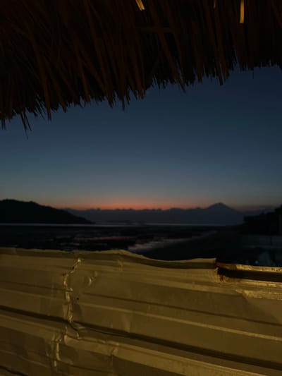 The image depicts a twilight scene overlooking a body of water with a silhouetted mountainous landscape in the distance. The foreground is dominated by a thatched roof and a corrugated metal structure, which appears to be some form of barrier or makeshift enclosure. The sky is a deep gradient of blue, transitioning to a faint band of orange and pink near the horizon, indicating the sun has recently set. The water's surface is dark and slightly reflective, with some indistinct shapes suggesting shallow areas or exposed seabed. The overall mood is tranquil and somewhat rustic, evoking a sense of being in a natural setting with man-made elements providing a partial enclosure. The location is identified as Kertasari, Indonesia, suggesting this is a coastal or lakeside view in that region.