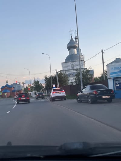 The image depicts a street scene in Bogorodskoe, Russia, likely during the daytime with soft lighting suggesting either early morning or late afternoon. The perspective is from a vehicle, looking out at the road and surrounding environment.
Dominating the left side of the frame is a large, white, multi-domed church with a silver roof. The church features architectural details like arched windows and intricate cross ornamentation on its spires. In the foreground, traffic is visible. A black sedan is parked to the right of the church, with a license plate that appears to read "X858BB 196". Further down the road, a line of cars is stopped at a traffic light. These include a white SUV with a license plate possibly ending in "B79". Another black vehicle is directly behind it, with a license plate ending in "3M". A red brick building with a blue dome, possibly another religious structure, is visible in the distance.
The road has clear lane markings. Electrical wires and streetlights are present, indicating an urban or suburban setting. Trees line the roadside, and some clouds are visible in the sky. The overall mood is calm and observational, capturing a typical moment in a Russian town. Visible text includes the license plates of the vehicles and some lettering on a building in the background that appears to be part of a water dispenser, with the word "ВОДА" (Water) partially visible.