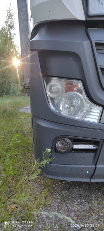 The image shows the front bumper and headlight assembly of a dark gray truck parked outdoors. The truck is positioned on the right side of the frame, angled towards the left. The background features a sun flare from the left, casting light through trees and onto tall green grass and a dirt road. The sky is bright, suggesting it is daytime. The scene appears to be in a rural or natural setting, possibly on the outskirts of Joutsa, Finland, given the mention of the location in the prompt. The truck's headlight is a complex unit with multiple lenses, and a fog light is visible below it. There are no people or discernible interactions in the image. The overall atmosphere is calm and natural.
