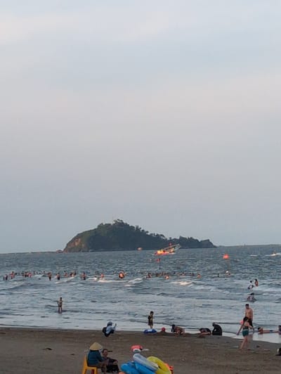 The image depicts a crowded beach scene in Hoằng Tiến Commune, Vietnam. The foreground is dominated by a sandy shore where several people are gathered. Two individuals are seated on chairs, with one wearing a conical hat, possibly engaged in conversation. Near them, a collection of inflatable water toys, including a blue and yellow ring, are visible. Further down the beach, several people are seen in various states of activity, some lying down, others crouching.

The middle ground of the image is occupied by the sea, filled with numerous bathers. The water is wavy, and many people are wading or swimming. A young boy stands in the shallow water, facing away from the camera.

In the background, a larger body of water stretches towards a prominent, verdant island. The island is covered in lush vegetation and appears to have some structures on its peak. A boat with visible lights is positioned in the water near the island. The sky above is overcast with a pale, light blue hue. The overall scene suggests a lively and popular recreational area.