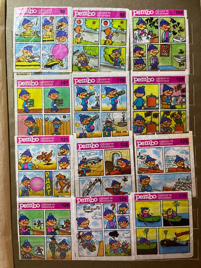 The image displays a collection of comic strip panels, likely trading cards or stickers, arranged in a grid. Each panel depicts a cartoon character, a young boy with a blue hat and scarf, engaged in various activities. The panels are organized into larger blocks, each with a pink header containing the word "pembo" and text that appears to be in Turkish: "çiğniyor ve düşünüyor" (chewing and thinking), followed by a number. The numbers visible are 88, 92, 100, 101, 119, 120, 121, 122, 125, 126, 129, and 130. The scenes within the panels range from playing with a bubble, carrying books, using a camera, being in a car, rowing a boat, and interacting with other cartoon characters and animals. The style is simplistic and humorous. The overall presentation suggests a product aimed at children. There is no indication of a specific location within Kramatorsk, Ukraine, or any urban environment depicted in the artwork. The background of the panels is varied, showing elements like grass, fences, interiors of rooms, water, and a snowy landscape. The image is a photograph of these collected panels, which are adhered to a backing material that appears to be cardboard or a similar surface. The lighting suggests an indoor setting with the photograph taken from a slight angle.