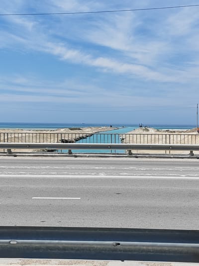 The image depicts a view from a road overlooking a bright blue channel of water, likely a canal, under construction. The water is a striking turquoise color and appears to be flowing into the sea in the distance. The area around the canal is sandy and appears to be undergoing significant earthmoving, with mounds of sand and construction equipment visible. 

In the foreground, a metal guardrail separates the road from the canal area. The road itself is asphalt with white lines marking lanes. The sky above is a clear blue with scattered white clouds, suggesting it is daytime. The overall scene is one of ongoing infrastructure development in a coastal or near-coastal environment. 

There are no people clearly visible in the image, but the presence of construction equipment indicates human activity related to the project. The weather appears to be good, likely sunny and warm given the clear sky and the context of outdoor construction. The location context of Ömirzaq, Kazakhstan, suggests a region where such large-scale water infrastructure projects might be undertaken, possibly related to land reclamation or transportation. No text is visible in the image.