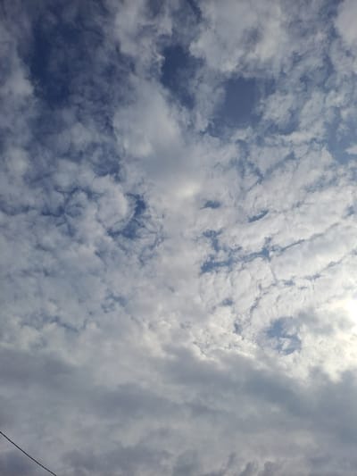 The image displays a daytime sky over the city of Avdot’ino, Russia, dominated by an arrangement of clouds against a blue background. White and light gray clouds, consistent with cumulus and stratocumulus formations, occupy the majority of the frame, varying in density and shape. Patches of clear, medium-blue sky are visible between the cloud masses, particularly in the upper left and central areas. The clouds in the lower and right portions of the frame appear denser and darker gray, suggesting varying thickness or lighting conditions, with a bright, possibly sunlit, area present towards the far right edge. A thin, dark, linear object, resembling an electrical wire or cable, extends diagonally upwards from the bottom-left corner of the frame, partially obscuring the lower sky and clouds. No individuals, explicit subjects, actions, or interactions are discernible.