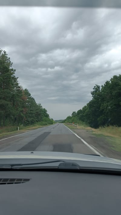 The image shows a view from inside a vehicle traveling on a two-lane asphalt road. The road is in moderate condition with some patched areas. Dense green trees line both sides of the road, creating a canopy effect in some sections. The sky is overcast with heavy gray clouds, suggesting a cloudy or possibly rainy day. The foreground shows the dashboard and windshield of the vehicle. In the distance, the road continues into a slightly hazy horizon. There are no people visible in the image. The scene appears to be a rural or semi-rural area.