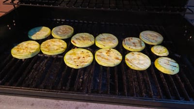 Sliced zucchini are grilling on a black metal barbecue in an outdoor setting. The zucchini slices are round with visible grill marks and a slightly charred appearance. The barbecue grate is dark and appears to be well-used. The background is dark, suggesting it is either nighttime or the area is shaded. The ground in the foreground is a rough, grey surface. The scene is located in Poletaevo 1th, Russia.