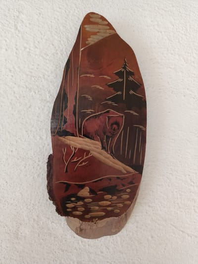 This is a carved piece of wood, likely a decorative plaque, depicting a woodland scene. The carving is done on a natural, irregularly shaped piece of wood, with the bark still visible on the left side. The carving features a bear in the center, standing on a hillside. Behind the bear, there is a pine tree and a stylized landscape suggesting mountains or hills in the background, with what appear to be clouds or a setting sun. In the foreground, there is a body of water with rounded shapes representing rocks or lily pads. The carving uses different shades of wood and possibly some dark staining to create depth and contrast. The overall impression is of a serene, natural environment. There is no visible text. The location context provided is Kürşım, Kazakhstan.