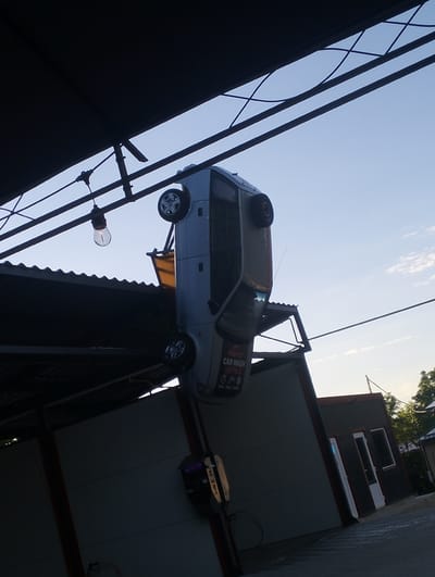 A silver car is suspended upside down as a decorative advertisement for a car wash. The car is positioned horizontally above a roofed structure, possibly part of the car wash facility. A single light bulb hangs from wires above the car. The sky is clear with a few scattered clouds. In the background, a building with windows and doors is visible, along with some trees and foliage. The image appears to be taken from ground level, looking upwards at an angle. The car wash is located in Șoldănești, Moldova.