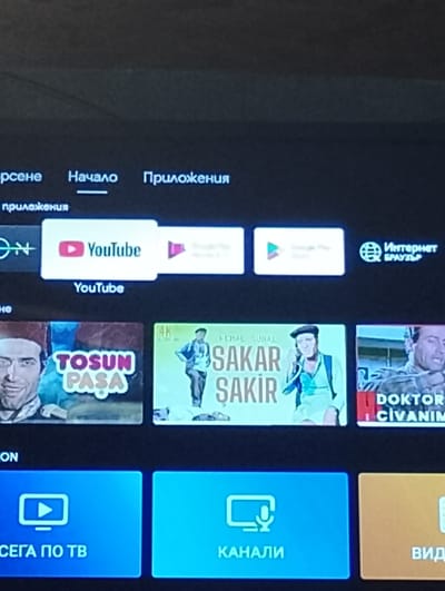 The image shows a television screen displaying a smart TV interface. The top of the screen features the words "Начало" and "Приложения," indicating "Home" and "Applications" in a Slavic language, possibly Bulgarian. Below, there are app icons, including YouTube. The next row down features movie posters for what appears to be Turkish comedies, namely "Tosun Paşa", "Sakar Şakir" and "Doktor Civanım". The bottom row shows icons and labels for "СЕГА ПО ТВ" ("NOW ON TV"), "КАНАЛИ" ("CHANNELS"), and a third option partially visible. It appears to be a snapshot of a home entertainment system interface. There is no visible indication of the city of Chernik, Bulgaria.