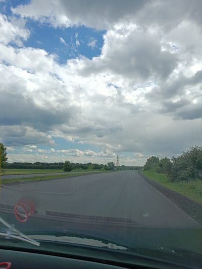 The image captures a view from a vehicle dashboard, looking out onto a two-lane asphalt road. The road appears to be in rural or suburban surroundings, with lush green fields and trees lining the sides. The sky is dramatic, filled with a mix of bright white and dark grey clouds, suggesting the possibility of impending weather changes. In the distance, a tall transmission tower is visible, hinting at the presence of infrastructure in the area. The dashboard itself is visible in the foreground, with a red coiled object and windshield wipers. The scene is likely captured while traveling on a road in or near the village of Biloziria, Ukraine.