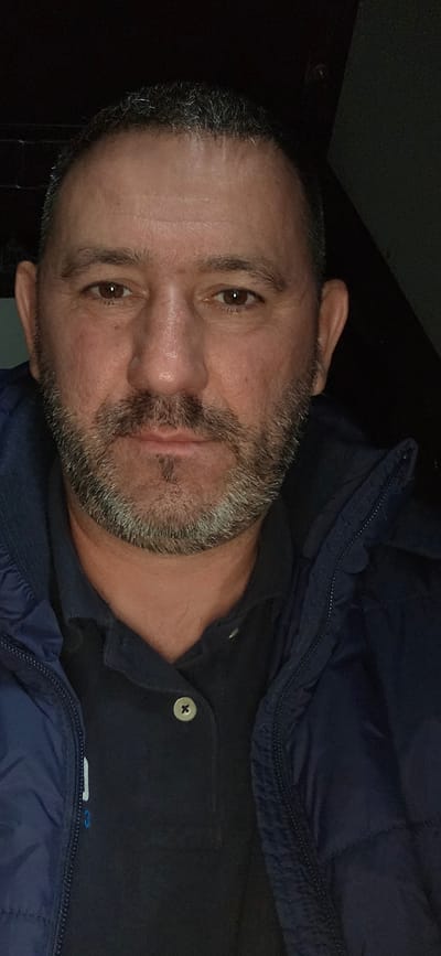 A close-up portrait features a man, likely in his mid-40s to 50s, looking directly at the viewer with a neutral expression. His head is slightly tilted. He has short, dark hair and a salt-and-pepper beard and mustache, showing a mix of dark and gray hairs. His eyes are dark brown. He is wearing a dark blue or black collared shirt, possibly a polo, with a visible light-colored button on the placket. Over the shirt, he wears a dark blue puffer jacket with a visible zipper running along its edge. The lighting appears dim, suggesting an indoor setting or nighttime conditions, with the background mostly obscured in darkness. No specific location cues are visible beyond the man himself and his attire. There is no discernible text. The image captures him in a moment of direct engagement with the camera. The location context provided is Mouscron, Belgium.