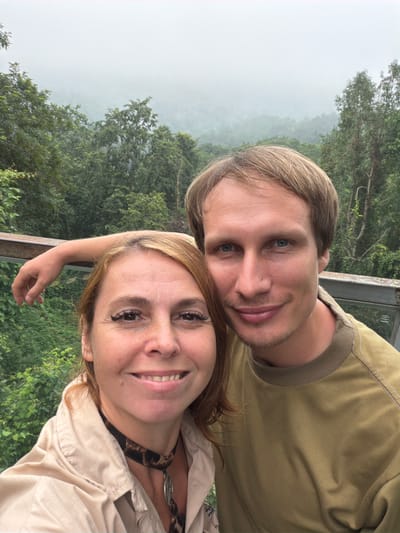 This is a selfie of a man and a woman taken outdoors with a misty, forested mountain landscape in the background, likely in Pong Yaeng, Thailand. The woman, on the left, has shoulder-length auburn hair and is wearing a light-colored jacket over a dark leopard-print top with a choker necklace. The man, on the right, has blond hair and is wearing a tan sweatshirt. Both are smiling at the camera, and the man's left arm is resting on a railing behind the woman. The environment appears to be a viewing platform or walkway amidst lush green trees, with fog obscuring the distant scenery.