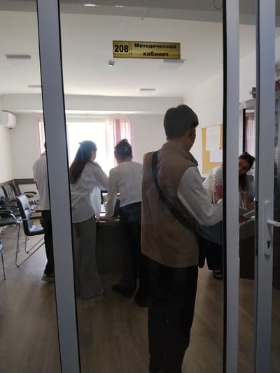 This image captures an indoor scene, likely an office or administrative building in Balykchy, Kyrgyzstan. The setting appears to be a reception or waiting area with chairs lined up against a wall and a desk in the background. A glass door, through which the photo is taken, reflects the interior.
Several people are visible. In the foreground, a person wearing a brown vest over a white shirt is facing away from the camera, towards the interior. They have a dark bag slung across their shoulder. Further inside, three individuals are interacting near a desk. One person, seen from the back, is wearing a white top and dark pants. Another person, also from the back, wears a white shirt and a long, beige skirt or trousers. A third person, partially obscured, is leaning over the desk. On the right side of the image, a woman with dark hair and glasses is looking towards the camera, peeking through the glass door.
The activity seems to be a casual interaction or a brief meeting at the desk. The room is brightly lit, suggesting it is daytime. The decor is simple, with a sign on the wall displaying "208" and Russian text that reads "Методический кабинет" (Methodical office/cabinet). The overall atmosphere seems neutral, with no strong indications of specific emotions. The floor is made of light-colored wood laminate.