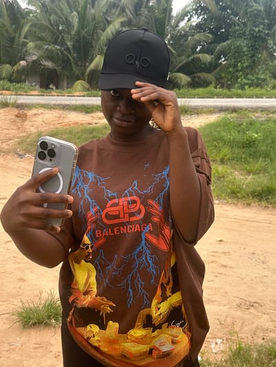 A young Black female is captured in a medium shot, standing outdoors in what appears to be a rural or semi-urban setting, possibly in Ganyingbo, Nigeria. She is wearing a brown t-shirt with a graphic print featuring abstract red and blue lightning bolts and a stylized image of a person wearing sunglasses. She also sports a black baseball cap with a logo. The woman holds a silver smartphone in her left hand, positioned as if taking a selfie, and her right hand is raised to shade her eyes from the sun. The background consists of lush green palm trees and vegetation, with a dusty path or road visible behind her. The lighting suggests it is daytime, with soft, diffused light.
