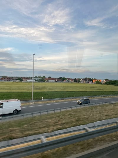 The image shows a view from a moving vehicle on a highway, possibly in the Surcin Urban Municipality, Serbia. On the right side of the highway, there is a grassy area followed by a town with houses and trees. A white van and a dark sedan are traveling on the highway. The sky is visible with scattered clouds, and the image appears to be taken during daylight. The image is captured through a window, which introduces a slight reflection on the left side.