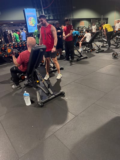 A gym setting features multiple individuals using exercise equipment. Several people are lifting dumbbells, while others use weight benches. A bottle of water rests on the floor beside one bench. A screen in the background displays an image of food. The gym appears busy with multiple machines in use.