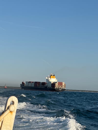 A large container ship, identified as "MARLA BULL" by the text on its stern, sails on the Adriatic Sea near Ravenna, Italy. The ship is laden with numerous red and white shipping containers stacked high. In the foreground, waves churn and break, with the bow of another vessel visible. A smaller red tugboat is seen to the left of the container ship, and a hazy shoreline with trees and structures is in the distance. The sky is clear and blue with faint contrails. The scene appears to be a sunny day with moderate sea conditions.
