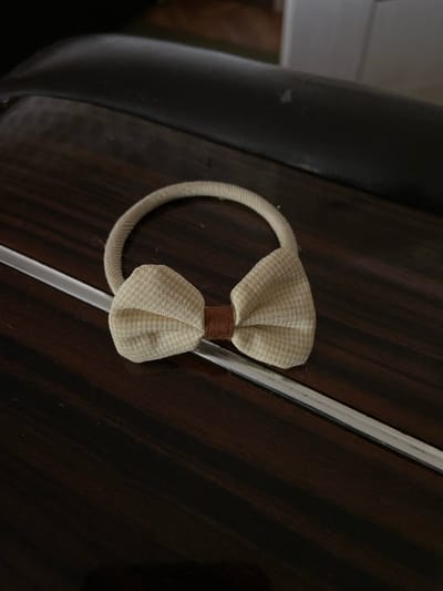 A beige and brown fabric hair tie with a bow is placed on a dark wood surface. A clear plastic strip runs diagonally across the image, beneath the hair tie. The background is dark and out of focus, with hints of dark furniture and possibly a white cabinet. The image is taken in Kojomkul, Kyrgyzstan.